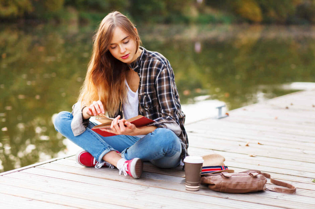 teen girl reading a book on a pier on the lake